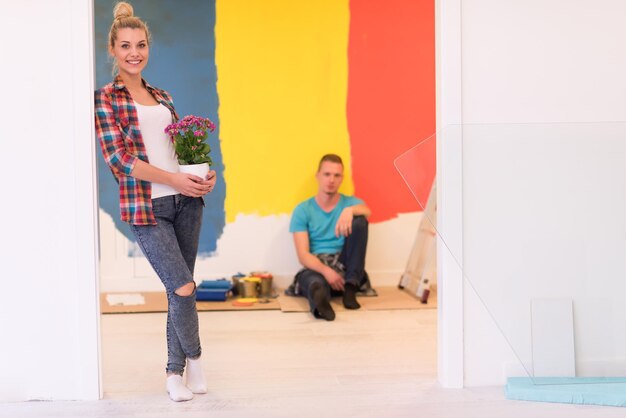 Happy couple doing home renovations, the man is painting the room and the woman hold the pot with flowers