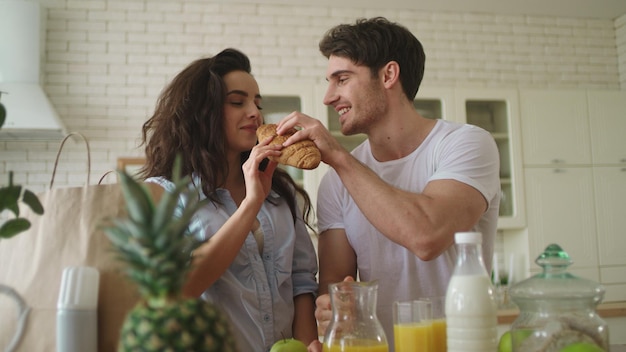 Photo happy couple eating croissant at kitchen joyful family playing with food