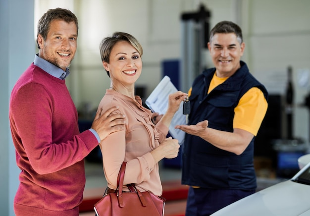 Happy couple giving car key to their auto repairman while looking at camera in a repair shop