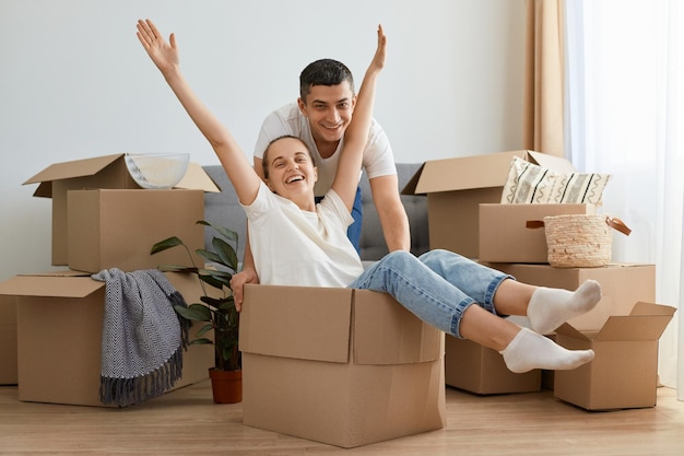 Happy couple having fun while moving into new house excited woman with raised arms riding sitting in cardboard box while man pushing it cheerful family playing while unpacking belongings together