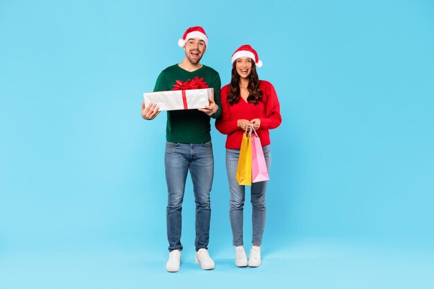 Happy couple holding shopper bags and wrapped present blue background