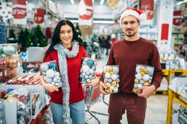 幸せなカップルは、店でクリスマスのおもちゃ、家族の伝統の箱を持っています。 12月のホリデーグッズやデコレーションの買い物