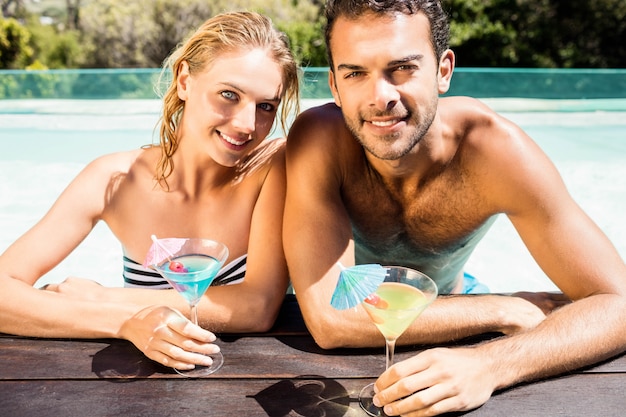 Happy couple leaning on pool edge and holding cocktails in a sunny day