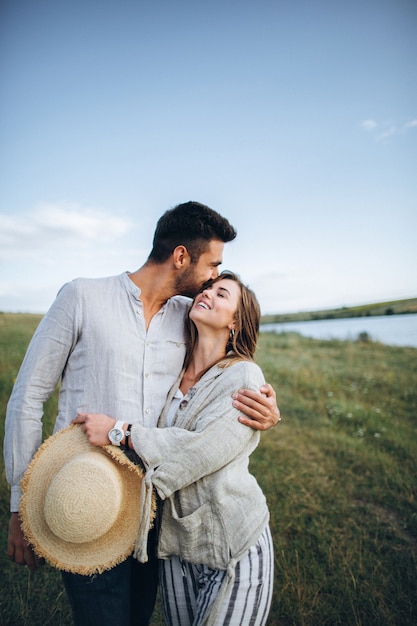 Foto coppia felice innamorata che abbraccia, bacia e sorride contro il cielo in campo. cappello in mano della ragazza