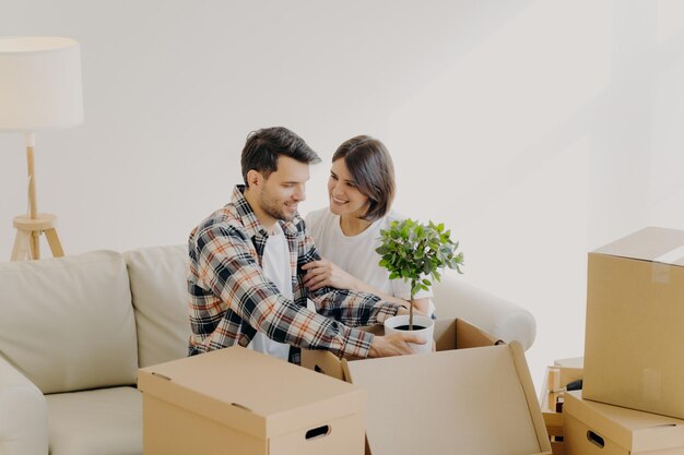 Happy couple in love settle in new home unpack boxes with household objects man holds potted plant woman embraces and expresses love to husband start new family life in modern apartment