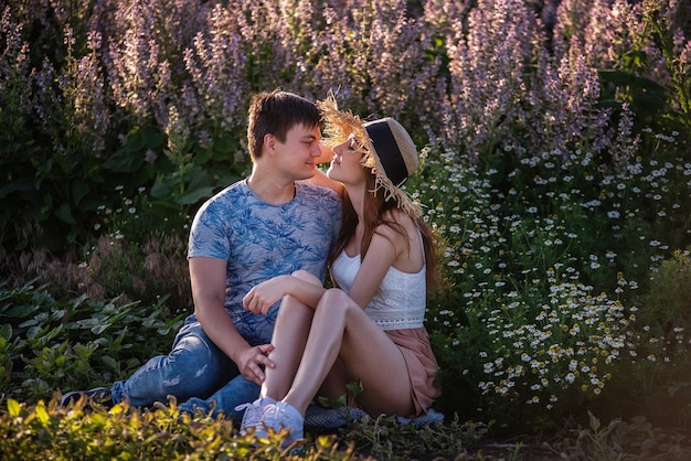 Happy couple in love sits hugging by a blooming sage field. A young man passionately embraces, holds hands, looks into the eyes of a beautiful girl in a straw hat.