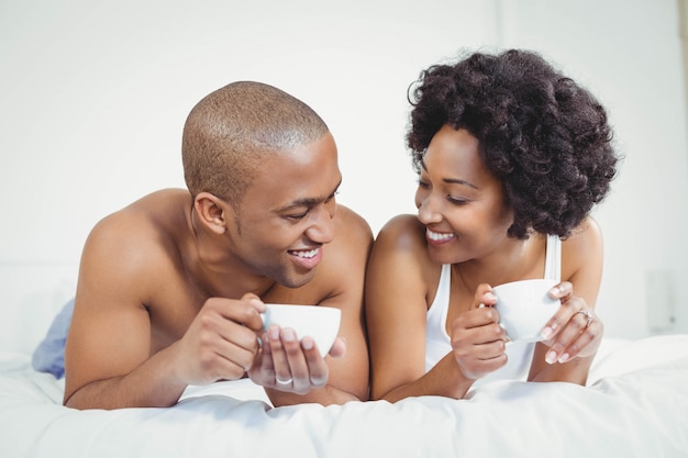 Happy couple lying on bed while holding cups and talking