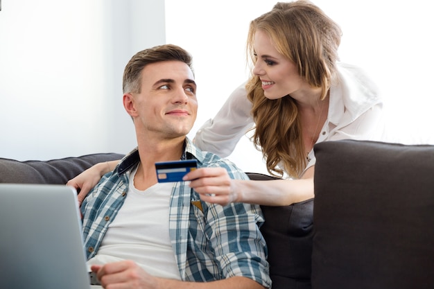 Happy couple making purchases in the internet using laptop and  credit card sitting on sofa in living room