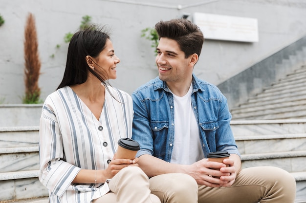 happy couple man and woman in casual clothes drinking takeaway coffee while sitting on bench near stairs outdoors