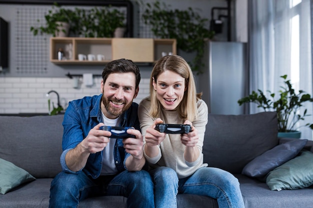 Happy couple man and woman playing game using joystick while sitting on sofa at home