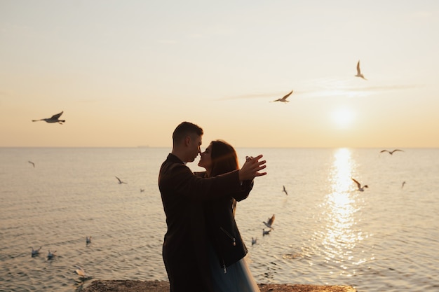 Happy couple of man and woman raised their hands up on the background of sunset and flying seagulls at sea.