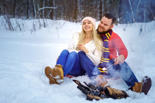 Happy couple man and woman in winter in the forest fry marshmallows on the fire romantic picnic