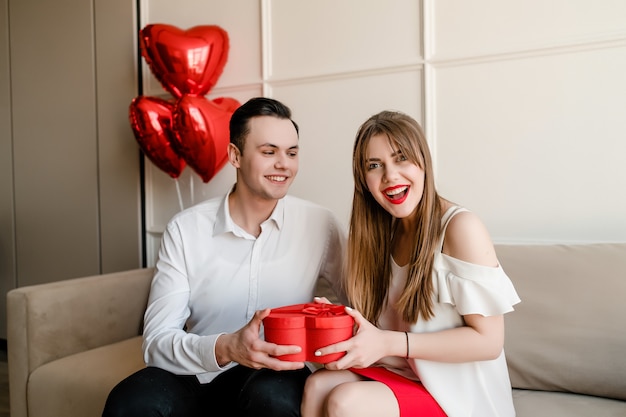 Happy couple man and woman with heart shaped red gift box and balloons on couch at home