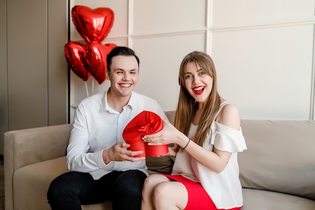 Happy couple man and woman with heart shaped red gift box and balloons on couch at home