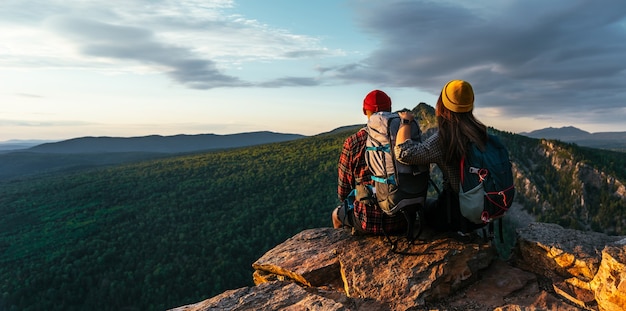 A happy couple in the mountains admires the beautiful views. A man and a woman with backpacks on the mountain admire the panoramic view. Travelers enjoy climbing the mountain at sunset. Copy space