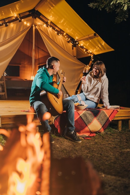 Happy couple relaxing in glamping on autumn evening drinking\
wine and playing guitar near cozy bonfire luxury camping tent for\
outdoor recreation and recreation lifestyle concept