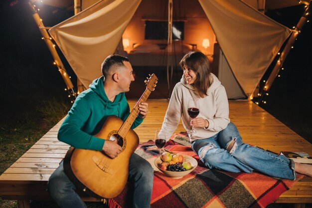 Happy couple relaxing in glamping on autumn evening drinking
wine and playing guitar near cozy bonfire luxury camping tent for
outdoor recreation and recreation lifestyle concept