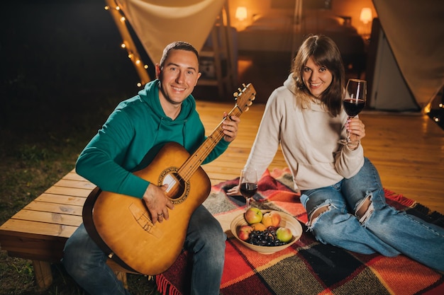 Happy couple relaxing in glamping on autumn evening drinking\
wine and playing guitar near cozy bonfire luxury camping tent for\
outdoor recreation and recreation lifestyle concept