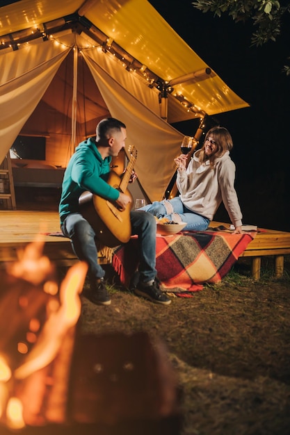 행복한 커플은 가을 저녁에 글램핑을 즐기며 아늑한 모닥불 근처에서 기타 연주와 와인을 마시고 야외 레크리에이션 및 레크리에이션 라이프스타일 개념을 위한 럭셔리 캠핑 텐트