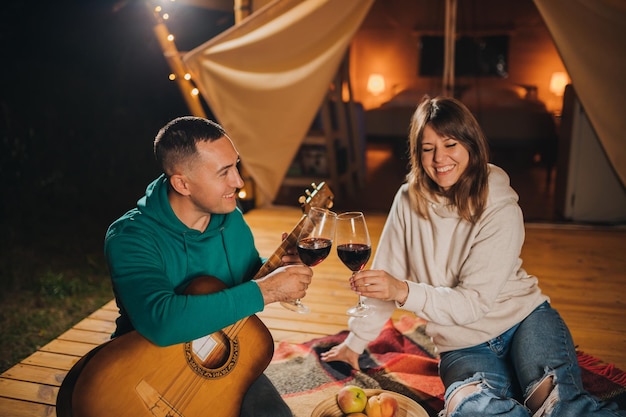 Coppie felici che si rilassano nel glamping la sera autunnale bevendo vino e suonando la chitarra vicino al falò accogliente tenda da campeggio di lusso per attività ricreative e ricreative all'aperto concetto di stile di vita