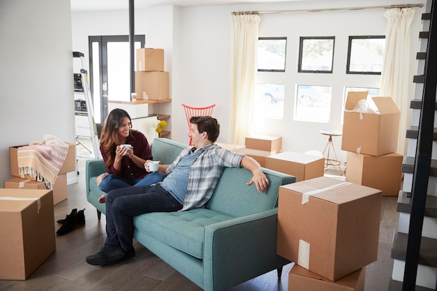 Happy Couple Resting On Sofa Surrounded By Boxes In New Home On Moving Day