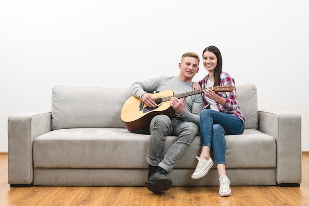 The happy couple sit on a sofa and play the guitar on the white wall background