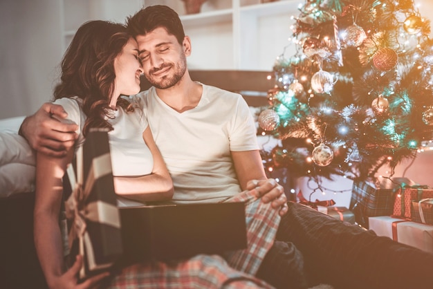 幸せなカップルは、クリスマスツリーの近くにギフトボックスを持って座っています