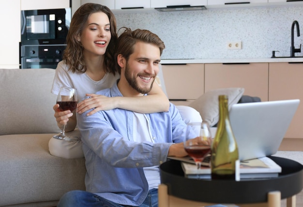 Счастливая пара сидит, отдыхая на полу в гостиной, используя ноутбук для покупок в Интернете, вместе смотрит фильм
