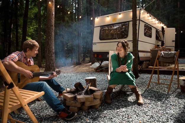 Happy couple smiling woman and man playing guitar sits close to campfire and trailer