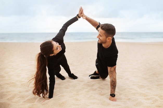 Happy couple in sportswear standing in side plank and giving high five training in morning on ocean