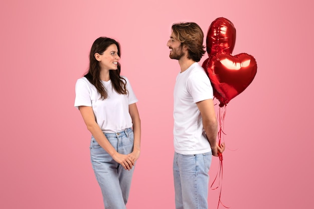 A happy couple stands facing each other with a playful expression holding shiny red heartshaped