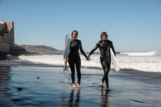 Coppia felice di surfisti camminando e ridendo lungo la riva del mare con sabbia nera in giornata di sole