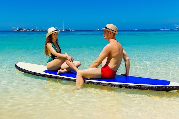 水着姿の幸せなカップルは、熱帯の海のパドルボードで楽しんでいます