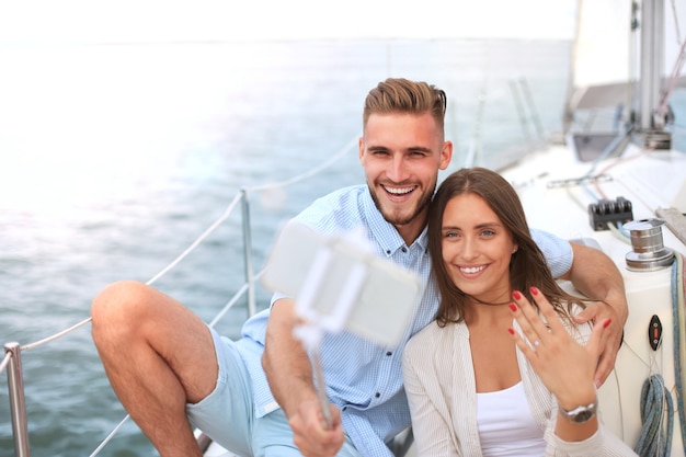 Coppia felice che si fa un selfie dopo la proposta di fidanzamento in barca a vela, rilassandosi su uno yacht al mare.