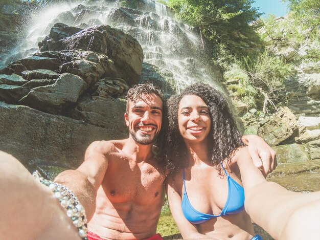Coppie felici che prendono il ritratto del selfie con la macchina fotografica dello smartphone sotto le cascate tropicali nelle vacanze estive