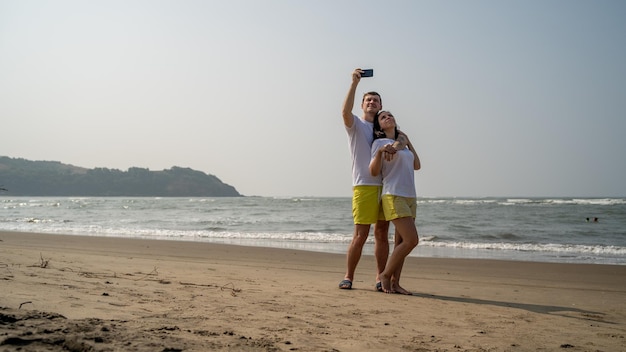 Happy couple taking selfies near sea Loving couple embracing during date on beach against waving sea and cloudless sky