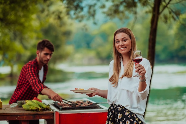 Happy couple toasting red wine glass while having picnic french dinner party outdoor during summer holiday vacation near the river at beautiful nature.