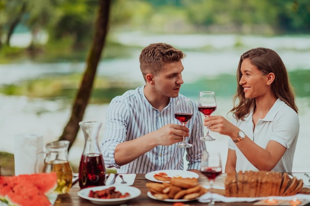 Happy couple toasting red wine glass while having picnic french dinner party outdoor during summer holiday vacation near the river at beautiful nature.