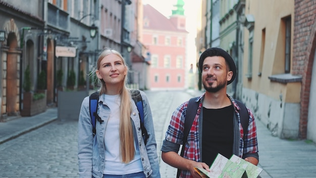 古いヨーロッパの都市の中央通りを歩いている地図を持つ観光客の幸せなカップル。彼らは周りを見回して笑っています。
