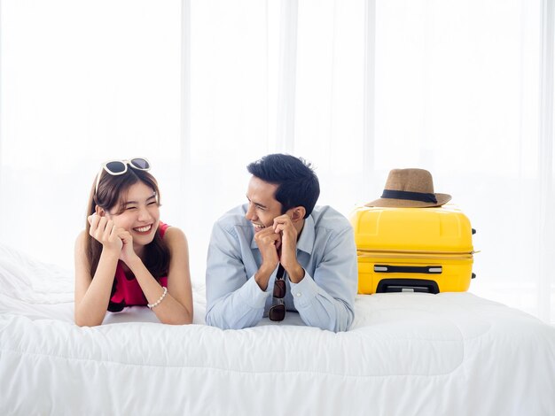 Happy couple travelers on honeymoon trip Asian man in denim shirt and young beautiful woman smile and lying together with yellow suitcases on bed in bedroom Happy holiday Colorful summer vacation