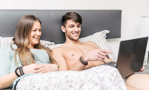 Happy couple watching things on computer, at home in the bedroom