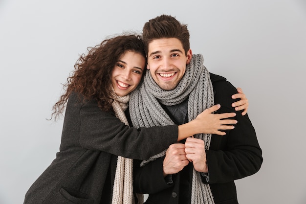 Happy couple wearing autumn clothes standing isolated over gray wall, hugging