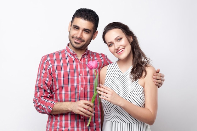Happy couple with tulip, smiling, hugging and looking at camera. Portrait of handsome man in red checkered shirt and beautiful woman in white striped dress standing. studio shot on grey background.