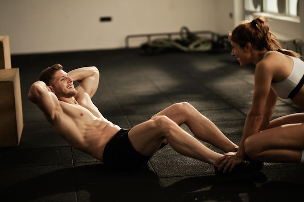 Happy couple working out in a gym together Man is doing situps while woman is assisting him