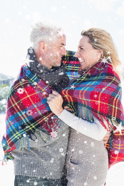 Happy couple wrapped up in blanket standing on the beach against snow falling