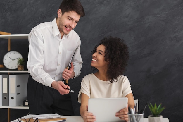 Happy couple of young business partners working in modern office. Two smiling coworkers discussing new project while working on digital tablet, copy space