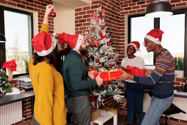 Happy coworkers exchanging festive presents and decorating christmas tree in office. Cheerful colleagues celebrating winter seasonal holiday at corporate party in coworking space