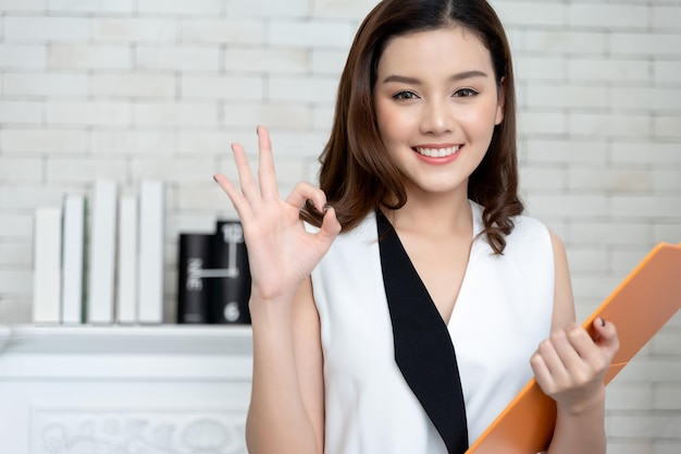 Happy creative young asian business woman in smart casual wear hand ok sign with holding clipboard looking to camera and happy smile modern office background