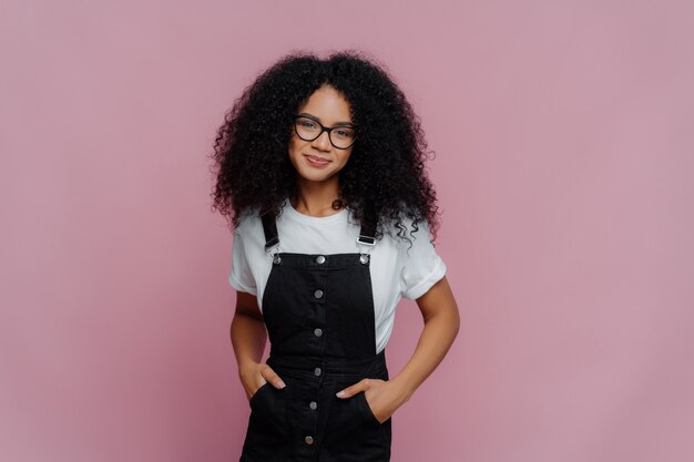 Happy curly African American woman keeps hands in pockets of sarafan and wears glasses