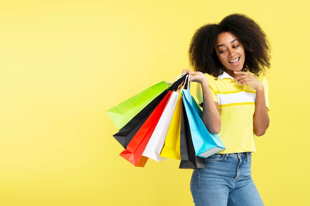 Happy curly haired African American woman holding colorful shopping bags isolated on yellow